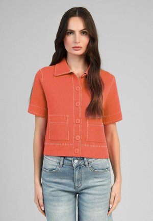 Femme aux longs cheveux foncés portant une chemise orange à manches courtes boutonnée avec coutures blanches et un jean bleu clair sur fond gris.