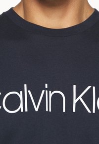 Granatowa bawełniana koszulka z krótkim rękawem z białym napisem "Calvin Klein" w wyraźnej, nowoczesnej czcionce na piersi.
