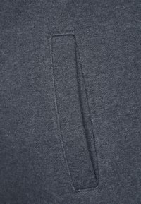 Tessuto di felpa grigio scuro con una sola tasca frontale discreta. La superficie è liscia con una leggera lucentezza. I bordi sono cuciti in modo ordinato.