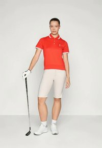 Sarkans polo krekls ar baltu apkakli un svītrām, gaiši brūni šorti, baltas sporta kurpes un golfa nūja, kas turēta labajā rokā.