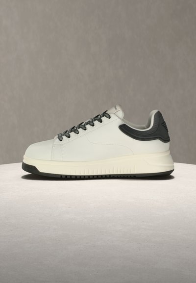 Λευκό αθλητικό παπούτσι με μαύρη σόλα και τακούνι, μαύρα κορδόνια και μαξιλαρωμένο γιακά, τοποθετημένο σε ανοιχτή επιφάνεια πάνω σε γκρι φόντο.