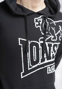 Sudadera negra con un estampado gráfico blanco de un león y el texto "Lonsdale". Tiene capucha con cordones y un tejido suave y texturizado.