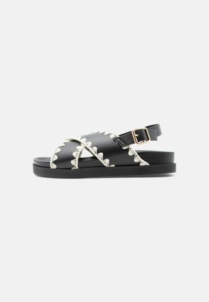 Sort flad sandal med hvide zigzag-syninger, krydsede remme og gyldent spænde på ankelrem, vist på hvid baggrund.