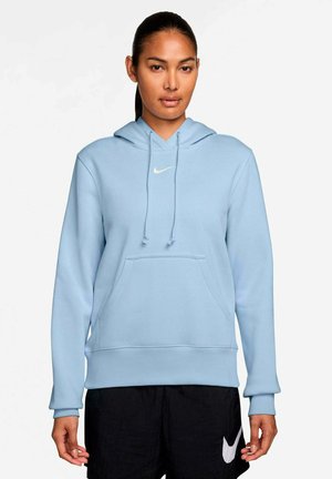 Frau trägt hellblauen Nike-Hoodie und schwarze Nike-Shorts, steht vor einem einfarbigen weißen Hintergrund.