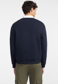 Sudadera de algodón de color navy con cuello redondo, con puños y dobladillo de canalé, que presenta un diseño minimalista. Lleva puesta sobre pantalones cargo verdes.