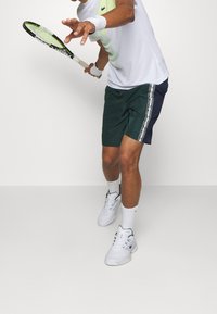 Tennis shorts i mörkgrön med marinblå detaljer, med ett sidobandmönster. Vita sportskor och en vit t-shirt med kort ärm bärs.