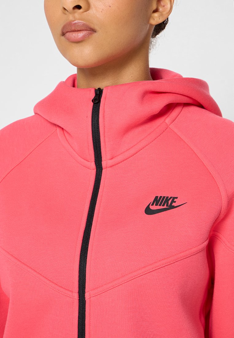 Sudadera con cremallera rosa y cremallera negra con el logo de Nike. Presenta una capucha y material texturizado con un diseño ajustado y costuras en contraste.