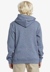 Modrá mikina s kapucí, která má strukturovaný povrch, žebrované manžety a lem. Disponuje stahovací kapucí a malou značkovou nálepkou na levém rukávu.