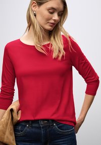 Rotes Langarmoberteil aus glattem Stoff mit Rundhalsausschnitt. Getragen über dunklen Jeans, mit Händen in der Taille. Minimalistisches Design, fokussierte Farbe.
