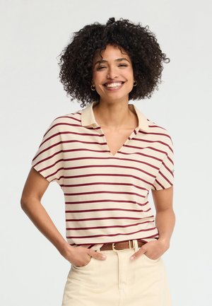 Uśmiechnięta kobieta z kręconymi włosami, ubrana w kremową i bordową koszulkę polo w paski z krótkim rękawem oraz beżowe spodnie, ręce w kieszeniach.