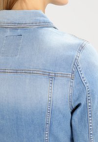 Jasnoniebieska kurtka dżinsowa z klasycznym kołnierzem, szwem na karczku z tyłu oraz kontrastującymi przeszyciami. Tkanina ma efekt postarzenia i teksturowaną powierzchnię.