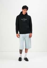 Joven vestido con una sudadera negra de Tommy Hilfiger, pantalones cortos de mezclilla azul claro hasta la rodilla, calcetines blancos y mocasines negros, de pie frente a un fondo blanco.