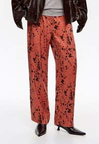 Pantalones de pierna ancha en un color óxido con patrones de manchas negras. Tela suave con una textura lisa, combinados con tacones negros puntiagudos.