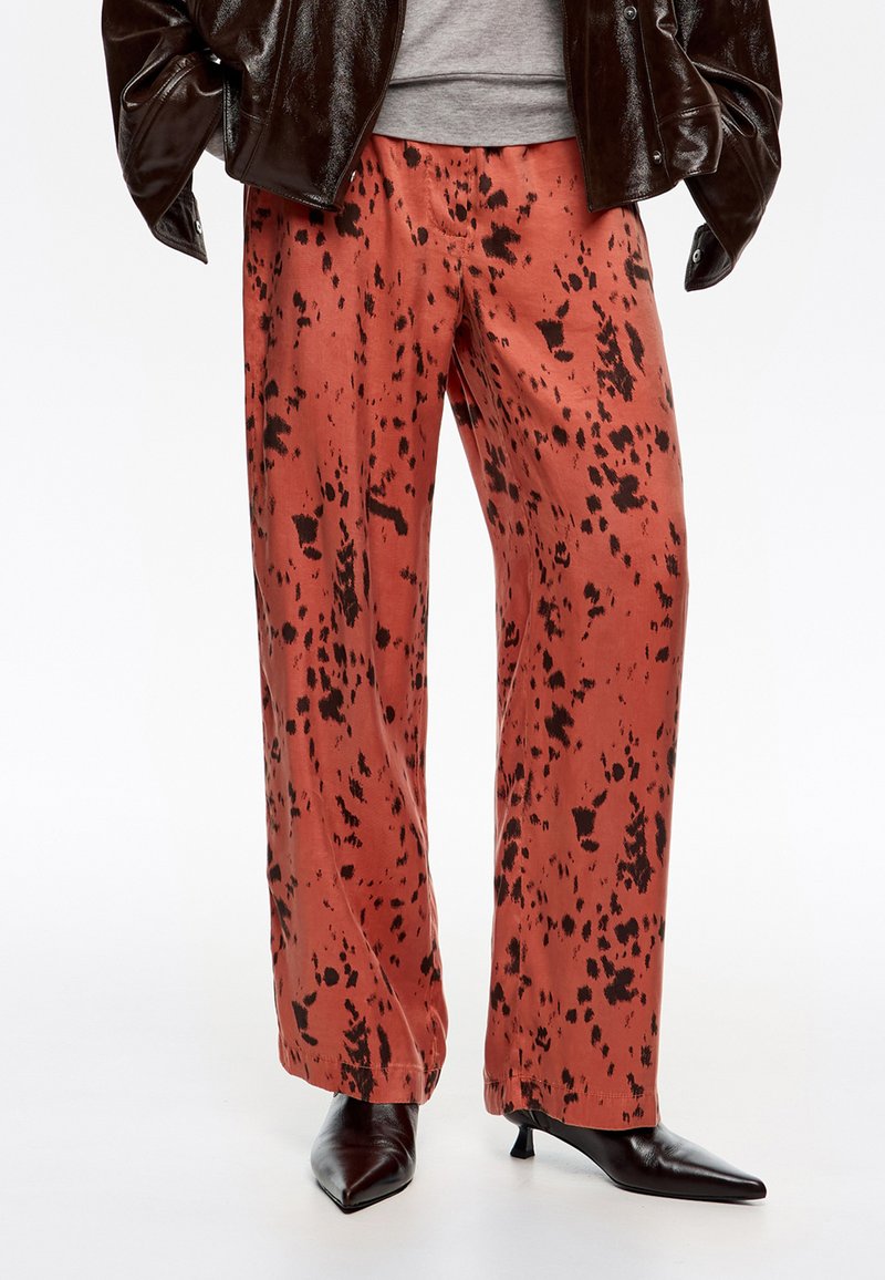 Pantalones de pierna ancha en un color óxido con patrones de manchas negras. Tela suave con una textura lisa, combinados con tacones negros puntiagudos.