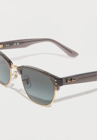 Γκρι και χρυσά γυαλιά ηλίου Ray-Ban με σκούρους φακούς και διαφανή μαξιλαράκια μύτης, τοποθετημένα σε λευκή επιφάνεια.