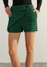 Shorts en tweed vert avec une finition texturée, présentant un design enroulé, des poches latérales et une fermeture à bouton à la taille.