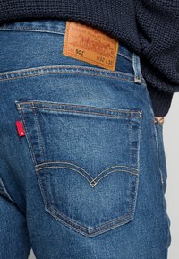 Denimjeans i medelblått med kontrasterande orange sömmar. Har en läderetikett märkt "501" och en liten röd logotyp på bakfickan.