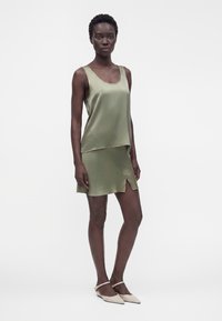 Femme debout portant un top en satin vert olive sans manches et une mini-jupe assortie avec une fente devant, associée à des ballerines blanches pointues.
