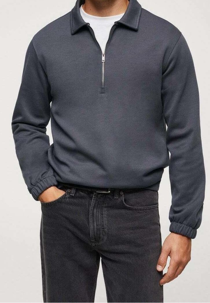 Pullover grigio con mezza cerniera, realizzato in tessuto morbido, caratterizzato da un colletto, polsini a coste e una texture sottile. Indossato sopra una camicia bianca e abbinato a jeans scuri.