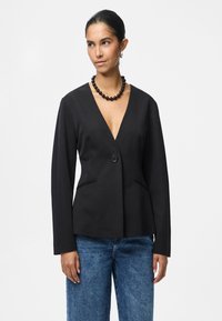 Černý sak na míru s hlubokým výstřihem do V, zapínání na jeden knoflík a dvě boční kapsy. Nosit přes modré džíny s vysokým pasem.