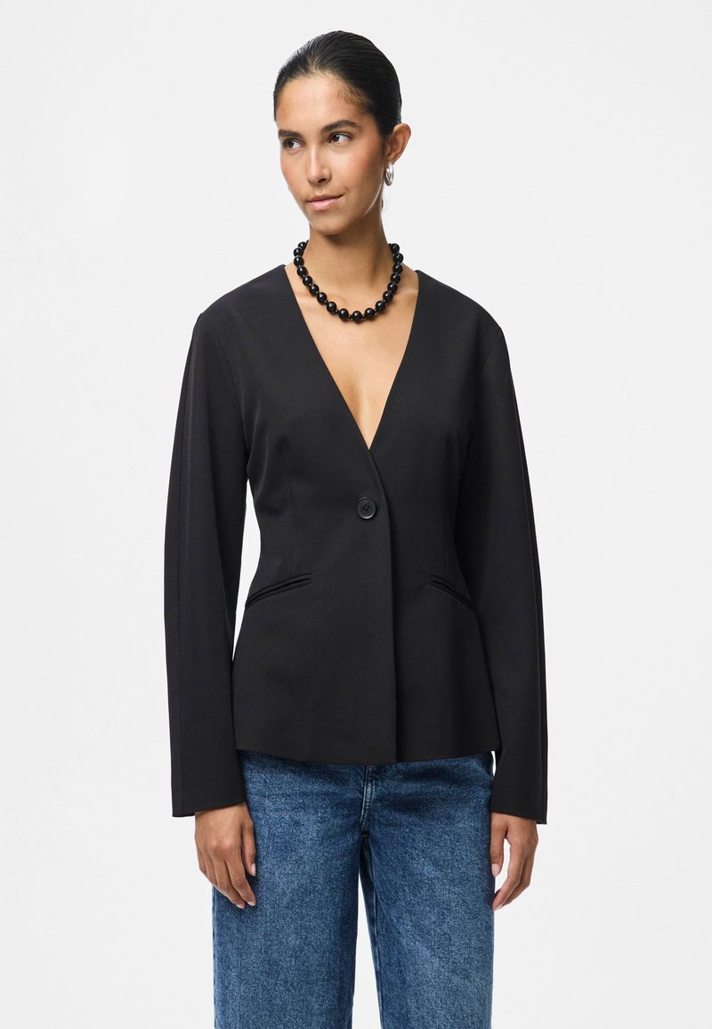 Černý sak na míru s hlubokým výstřihem do V, zapínání na jeden knoflík a dvě boční kapsy. Nosit přes modré džíny s vysokým pasem.