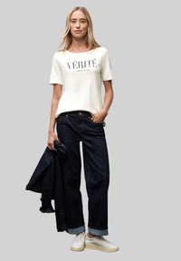 Hvid T-shirt med sort tekst, mørkeblå brede jeans og hvide sneakers. Den har en opsmøget kant og en jakke draperet over den ene arm.