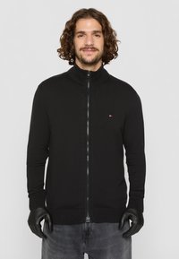Sweat à zip noir avec col montant, fabriqué en tissu tricoté texturé. Présente un petit logo sur la poitrine gauche et des poignets côtelés.
