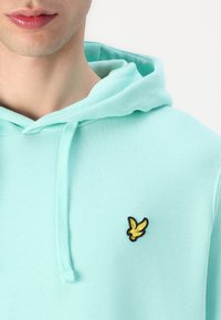 Svetlomodrá mikina s kapucňou a šnúrkami, s čiernym a žltým vyšitým logom na ľavej strane hrude. Hladká textúra látky.
