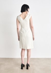 Sleeveless, V-neck cream dress with a fitted silhouette, back zipper, and knee-length hem. Features cap sleeves and a smooth texture.