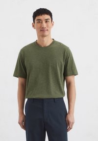 Green striped t-shirt with short sleeves, round neckline, and a relaxed fit. Worn with dark blue trousers against a plain background.