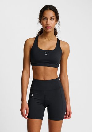 Mujer con cabello rizado que lleva un sujetador deportivo negro y pantalones cortos de ciclista negros de talle alto, de pie contra un fondo gris liso.