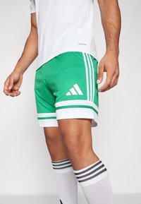 Zelené športové šortky s bielymi pruhmi a logom Adidas. Vyrobené z ľahkého materiálu, navrhnuté pre pohodlie a pohyb.
