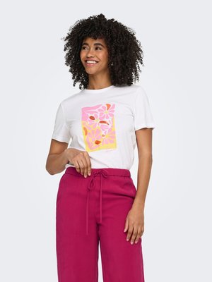 Femme souriante aux cheveux bouclés portant un t-shirt blanc à imprimé floral rose et jaune et un pantalon à cordon de serrage magenta, debout de manière décontractée.
