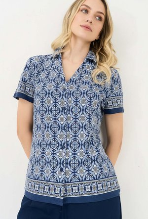 Femme blonde portant une chemise bleue à manches courtes avec des motifs floraux et géométriques, debout devant un fond clair uni.