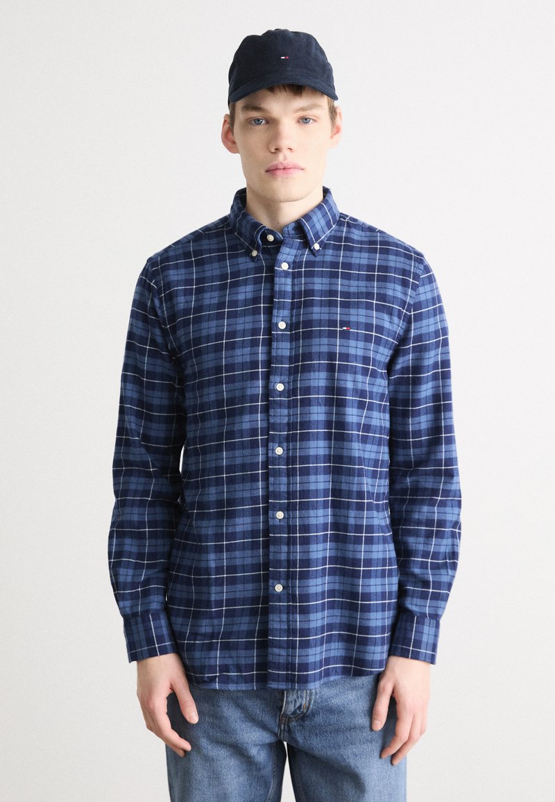 Camisa de cuadros azul marino con acentos blancos, de material de algodón, de mangas largas y con un pequeño logo. Combinada con una gorra oscura y jeans azules.