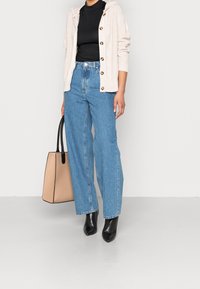 Jasno beżowy sweter z dzianiny z guzikami, czarna koszulka, niebieskie dżinsy z wysokim stanem oraz czarne botki za kostkę, trzymający beżową torbę na ramię z czarnymi akcentami.