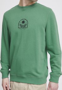 Felpa verde con maniche lunghe e scollatura rotonda. Presenta una stampa nera con un design di sole e vegetazione nella parte anteriore. Tessuto morbido al tatto.