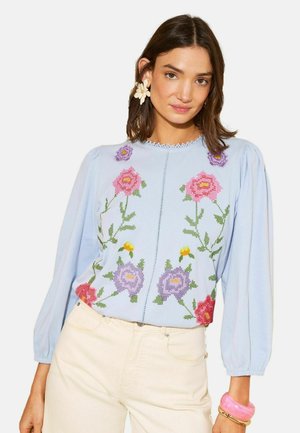 Femeie purtând o bluză albastru deschis cu broderie florală roz și mov, pantaloni crem, cercel în formă de floare și brățări roz și aurii.