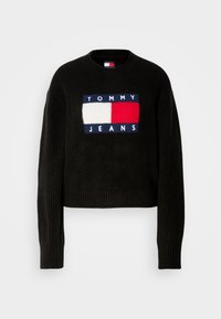 Svart croppet genser med ribbede ermer, med et strikket rødt, hvitt og blått logo-patch som sier "TOMMY JEANS" foran.