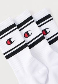 Trois chaussettes blanches montantes avec des rayures noires et un logo brodé rouge, noir et blanc près du bord supérieur.