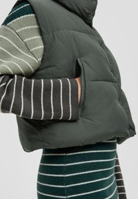 Grüne Steppweste mit hohem Kragen und Reißverschlusstaschen, getragen über einem gestreiften Pullover. Die Weste hat eine glatte Textur und ein gestepptes Design.
