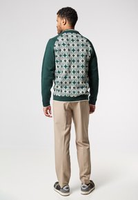 Pletený tmavě zelený kardigan s krémovým květinovým vzorem na zádech; žebrované manžety a límec, doplněno béžovými kalhotami a šedými teniskami.