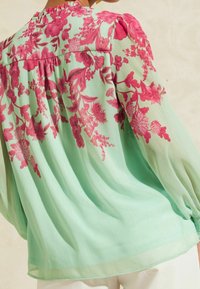 Blusa ligera en verde menta, con un audaz patrón floral en rosa. Mangas largas con detalles fruncidos en los hombros y la espalda.
