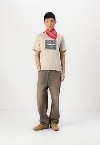 T-shirt beige clair avec un graphique carré gris, associé à un jean marron ample et des chaussures beiges. Un bandana rouge est noué autour du cou.