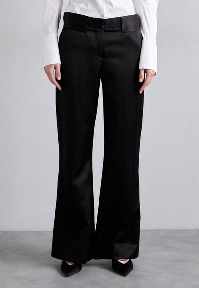 Schwarze, weit geschnittene Hosen aus einem glatten Stoff. Mit einem flachen Bund und Gürtelschlaufen. Kombiniert mit einem weißen Hemd mit Knopfleiste und spitzen Schuhen.