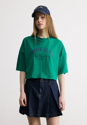 Giovane donna che indossa una maglietta corta verde Calvin Klein, gonna di jeans plissettata blu scuro e cappello blu navy, in piedi davanti a uno sfondo semplice.