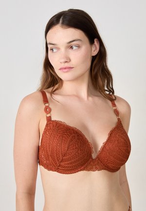 Model draagt een roestkleurige kanten bh, met ingewikkelde ontwerpen en verstelbare bandjes, terwijl zacht licht haar natuurlijke kenmerken accentueert.