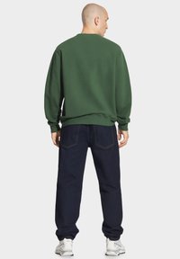 Zielona bluza z długim rękawem, z prążkowanymi mankietami i dolnym wykończeniem. Noszona z ciemnoniebieskimi dżinsami i białymi trampkami. Widok z tyłu.