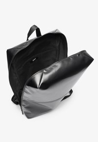 Mochila negra hecha de material brillante, con una forma angular, cierre de cremallera y un interior negro con una etiqueta. Bolsillo interior visible.
