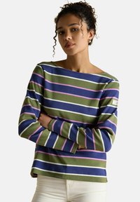 Kortsukli naise juuksed on lokkis, ta kannab pikki varrukatega särke, millel on horisontaalsed laevakujulised, oliivrohelised, roosad ja valged triibud, käed on ristis ja jalas on heledad püksid.